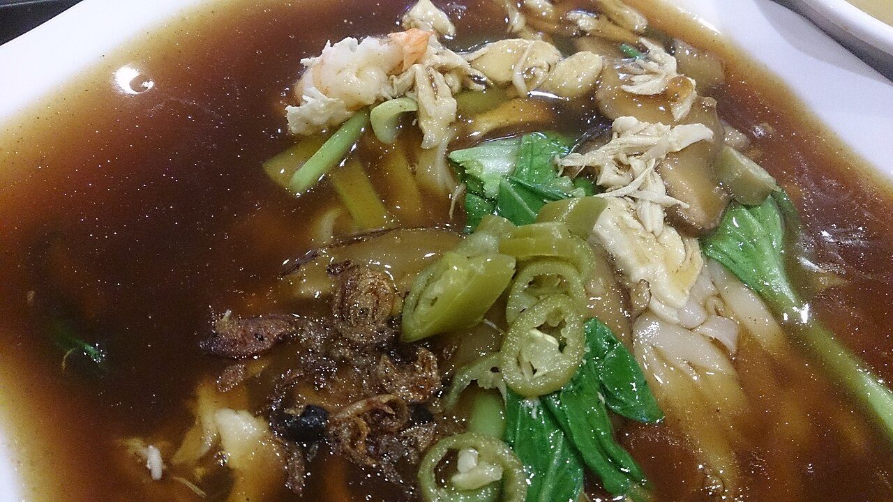 A bowl of Ipoh hor fun, flat rice noodles in dark gravy with prawn, chicken and greens, typical of Ipoh hawker food