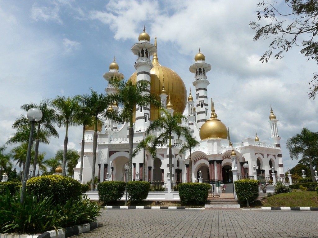 White Ubudiah Mosque with golden domes and minarets behind palm trees and a paved forecourt