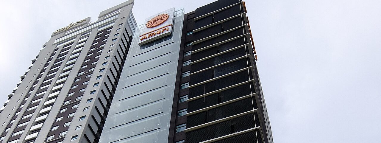 The upper floors of the Amari Johor Bahru hotel tower with its orange Amari sign, next to a taller residential tower, under a grey sky