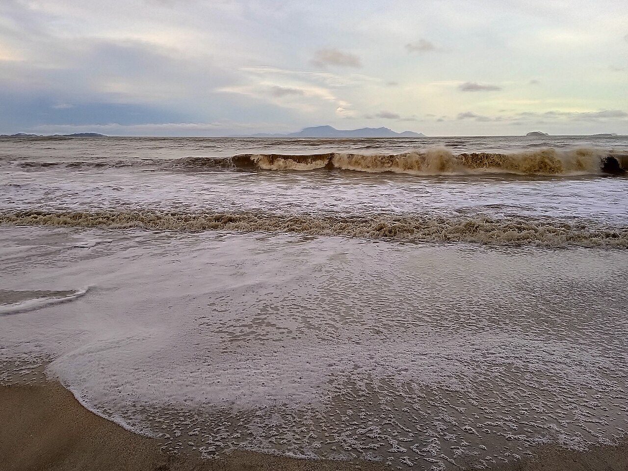 Brown, choppy waves breaking on the sand at Air Papan Beach near Mersing, with islands on the horizon