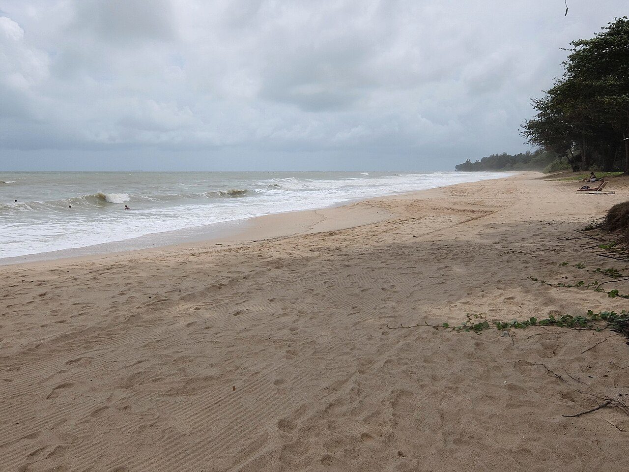 A wide, empty sandy beach at Desaru with breaking waves and casuarina trees under a cloudy sky