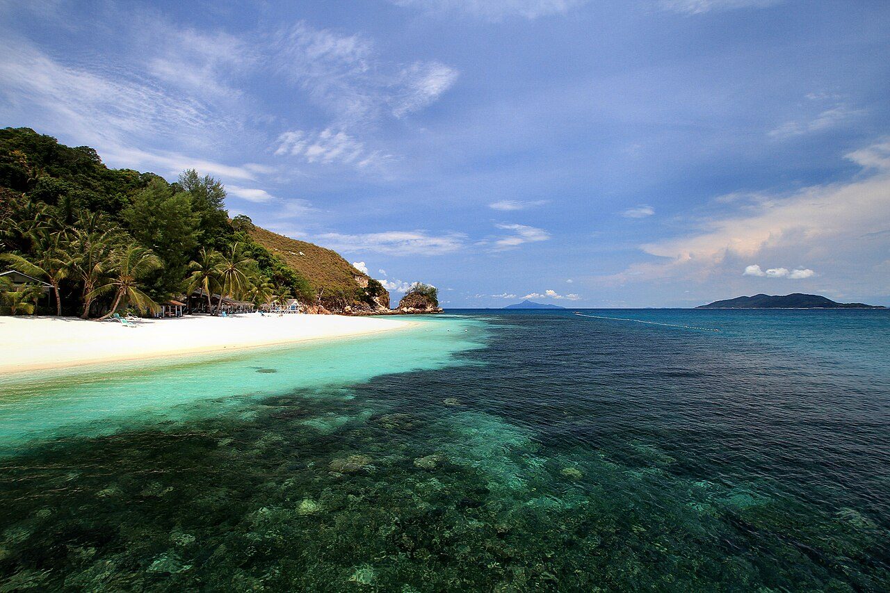 Coral patches in clear water off a white-sand beach with palms and a rocky headland at Pulau Rawa