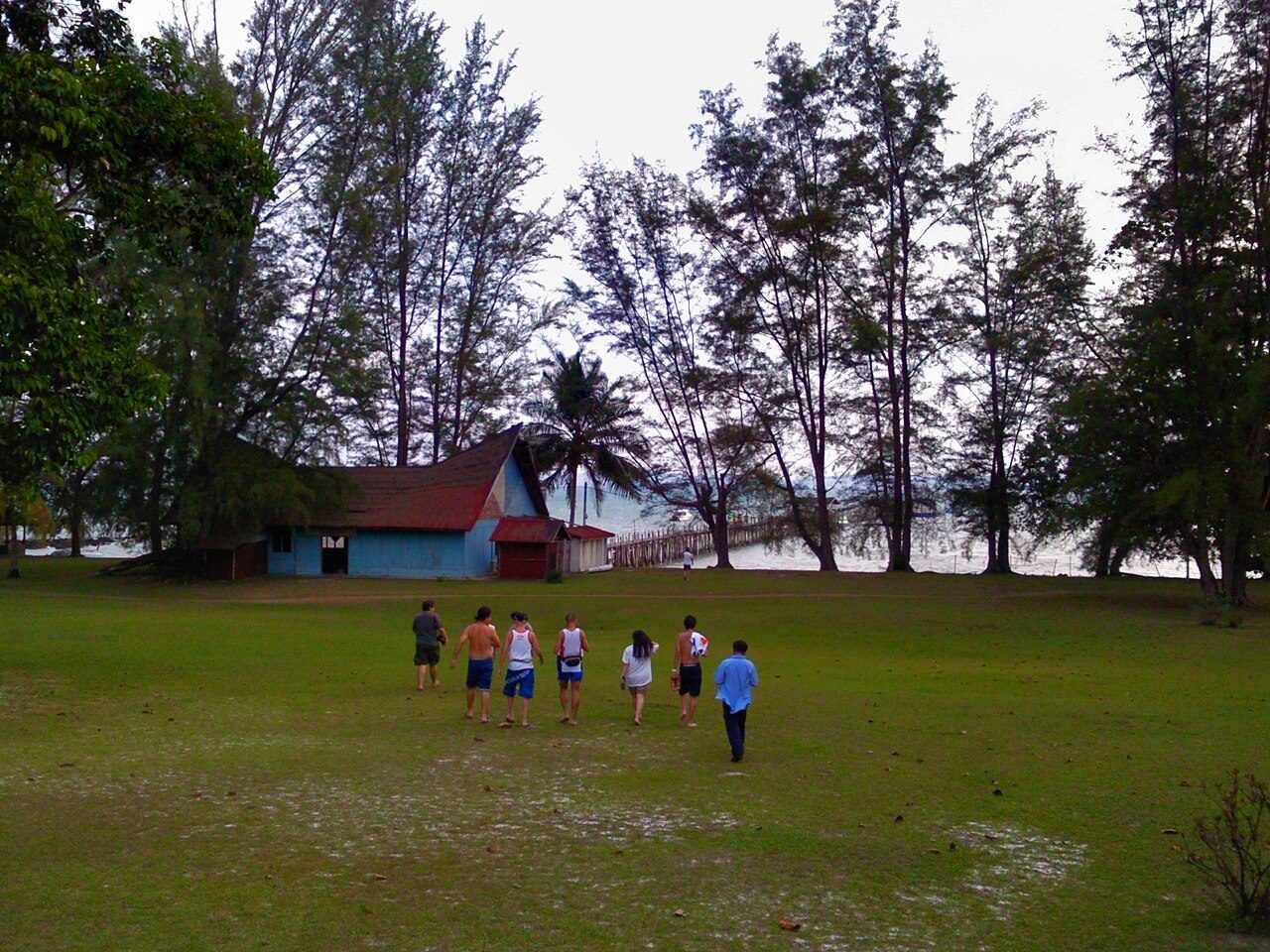 A group walking across a grassy clearing under casuarina trees towards a red-roofed building and jetty on Pulau Sibu