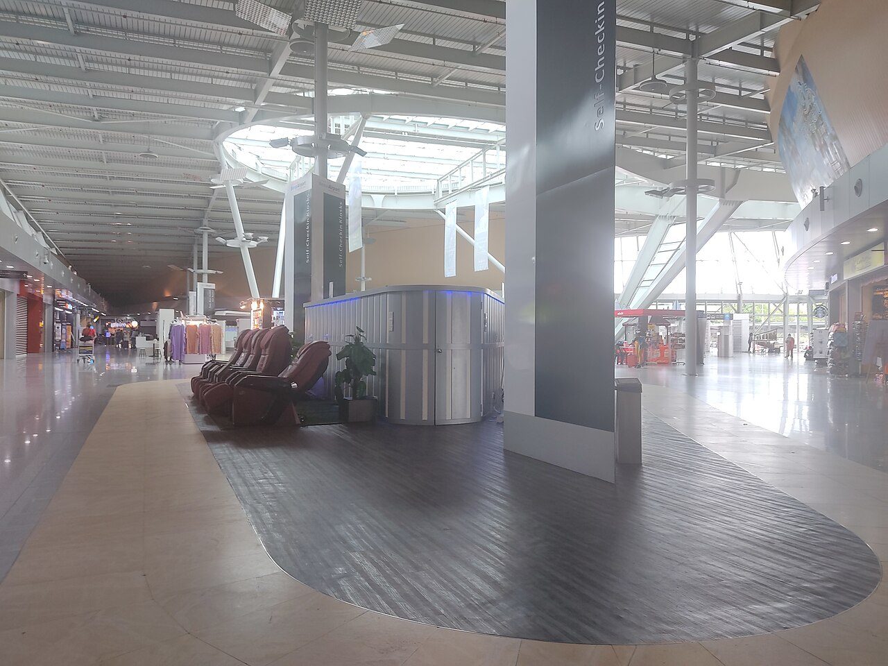 Inside the Senai Airport terminal: a long, bright concourse with massage chairs, a wooden floor panel and shops along one side