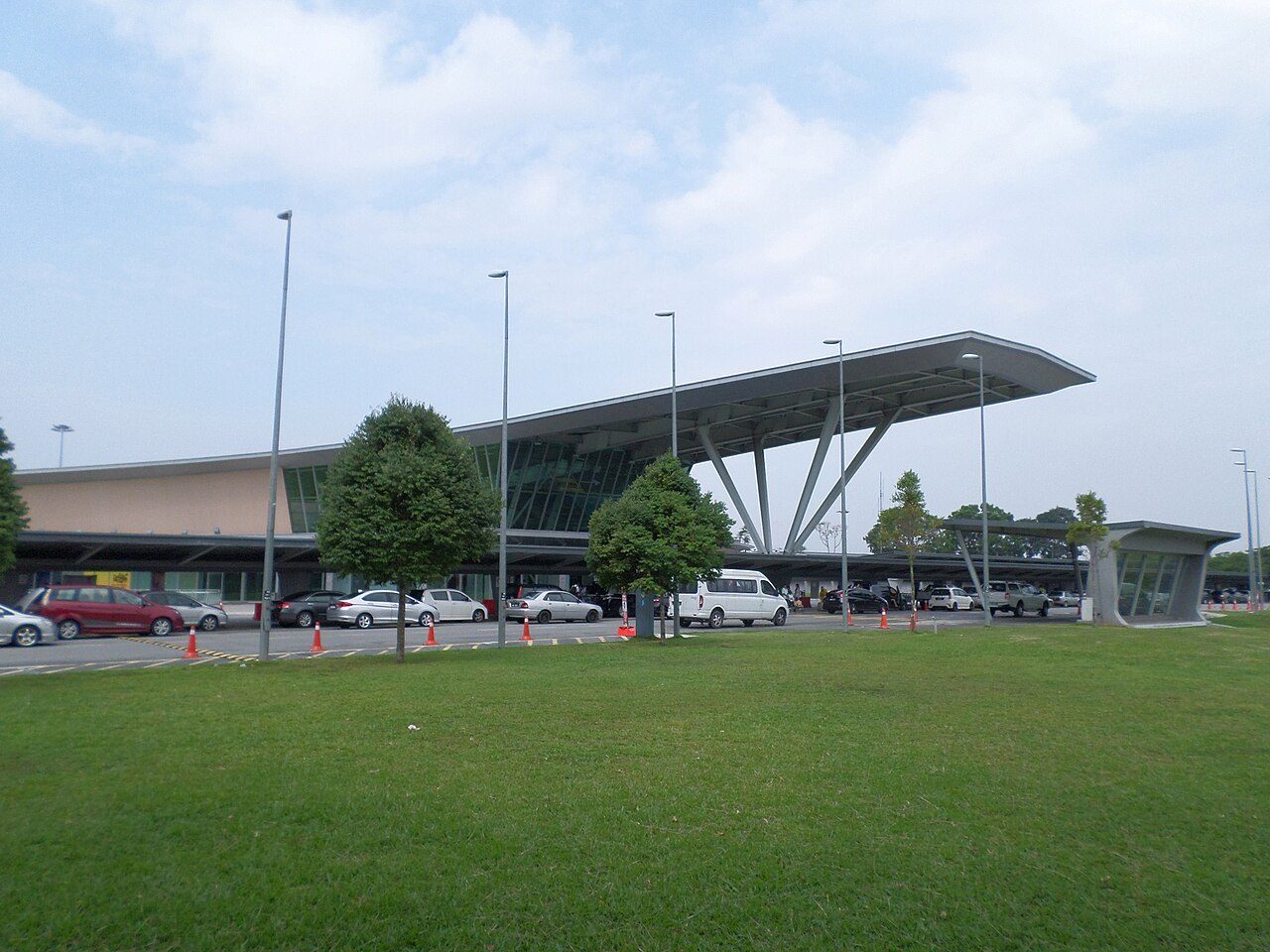 The front of the Senai International Airport terminal with its sloping white canopy, a row of parked cars and a lawn in the foreground