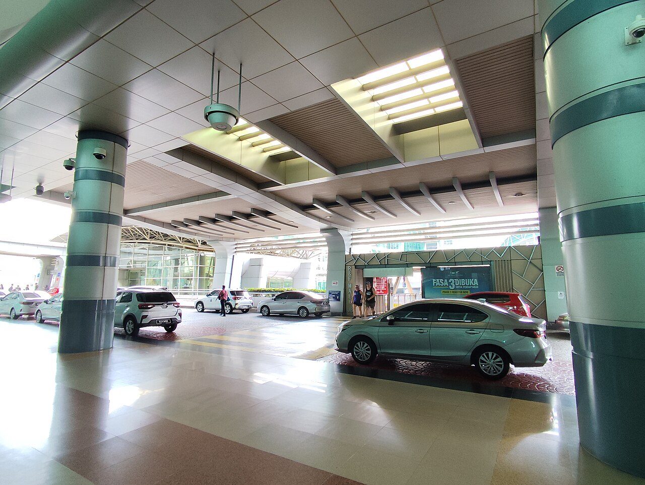 The covered pick-up point at JB Sentral station with cars waiting in the driveway and the link bridge entrance to R&amp;F Mall behind