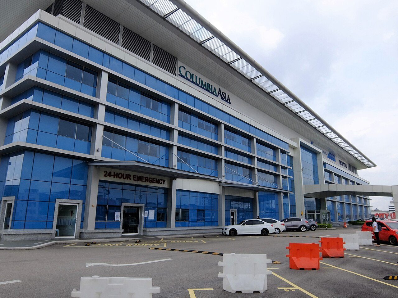 The blue glass front of Columbia Asia Hospital Tebrau, with a 24-Hour Emergency sign over the entrance and ambulance bays in front