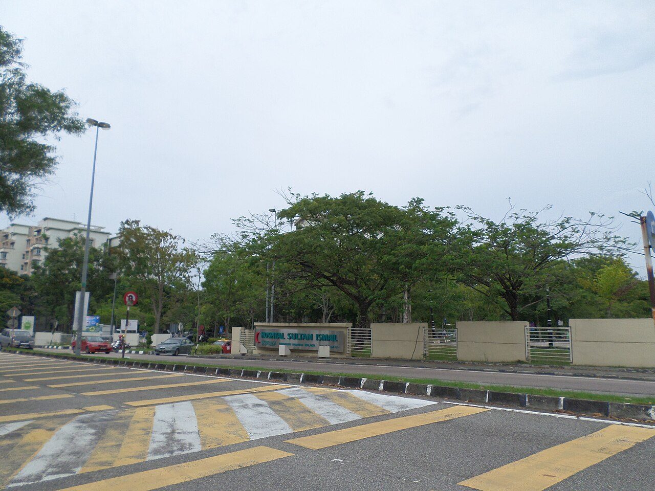 The roadside entrance sign of Hospital Sultan Ismail in Mount Austin, Johor Bahru, behind a row of large rain trees