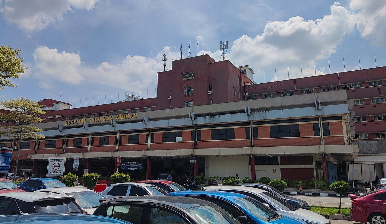 The front of Hospital Sultanah Aminah in Johor Bahru, a red-brick building with the hospital name in gold letters above a covered entrance, with cars parked in front