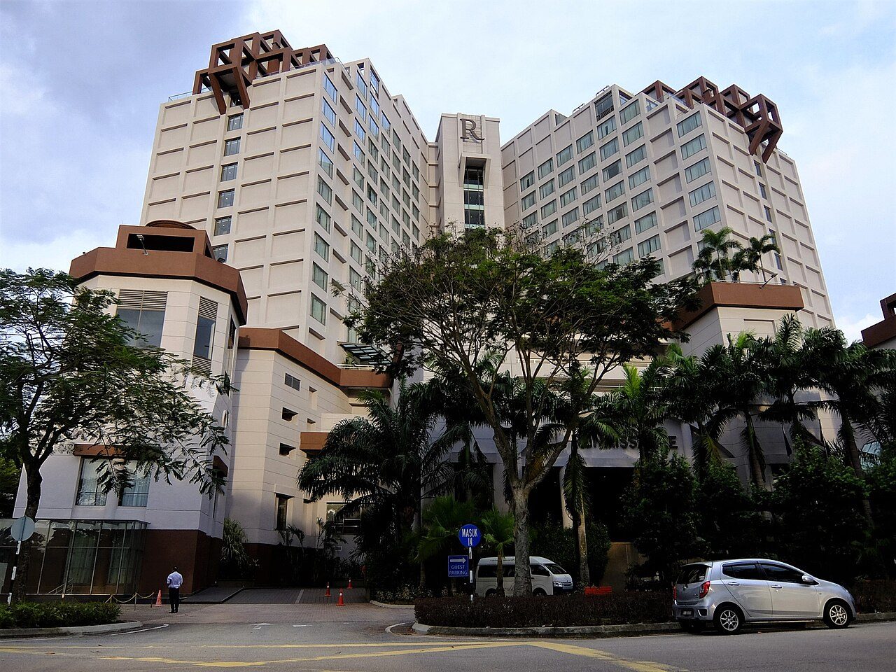 Renaissance Johor Bahru Hotel, two cream towers with brown lattice tops and the R logo, behind trees at the entrance in Permas Jaya