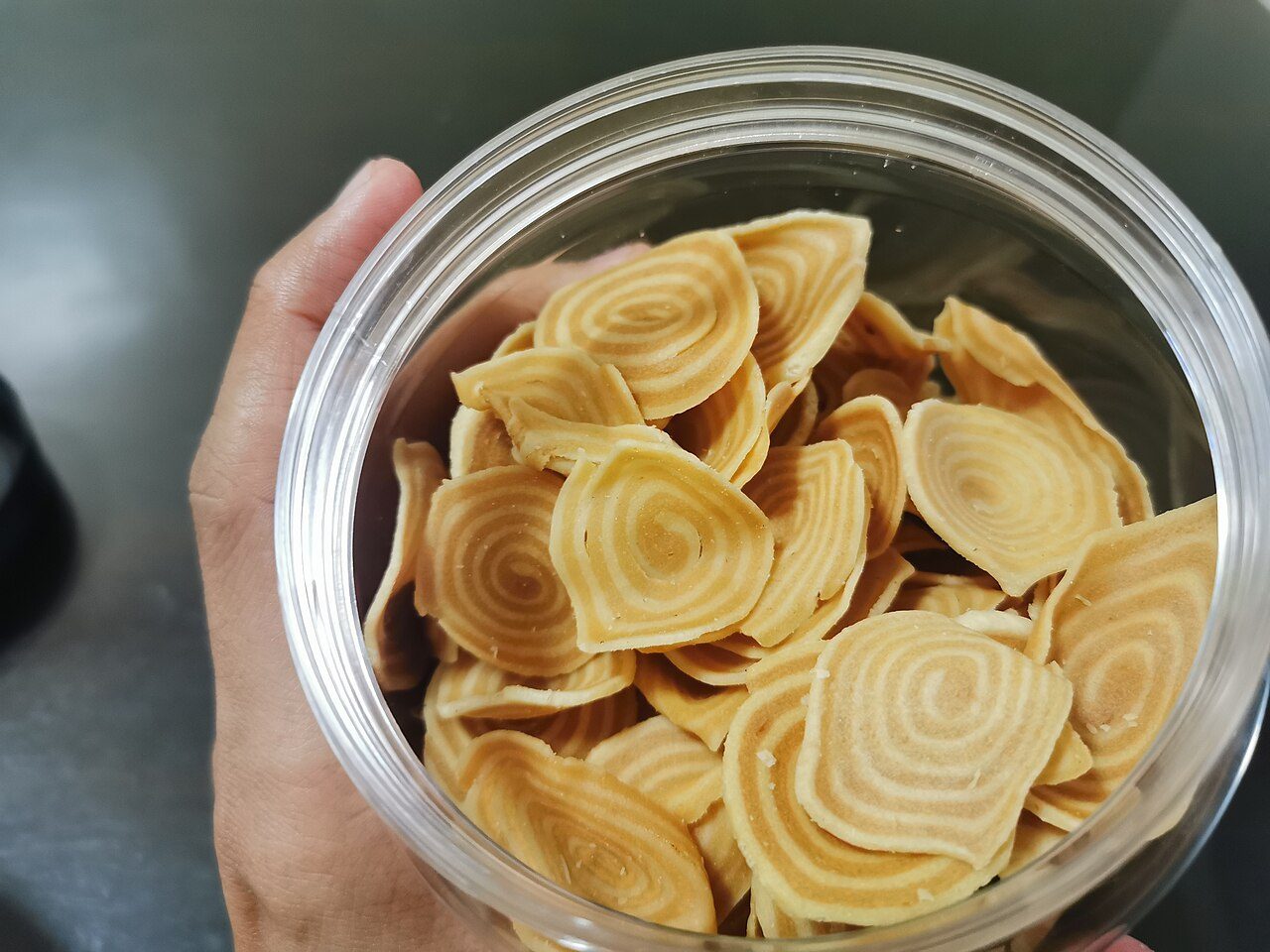 A hand holding a clear tub of kerepek bawang, thin spiral-shaped crackers sold in packets in Malaysia