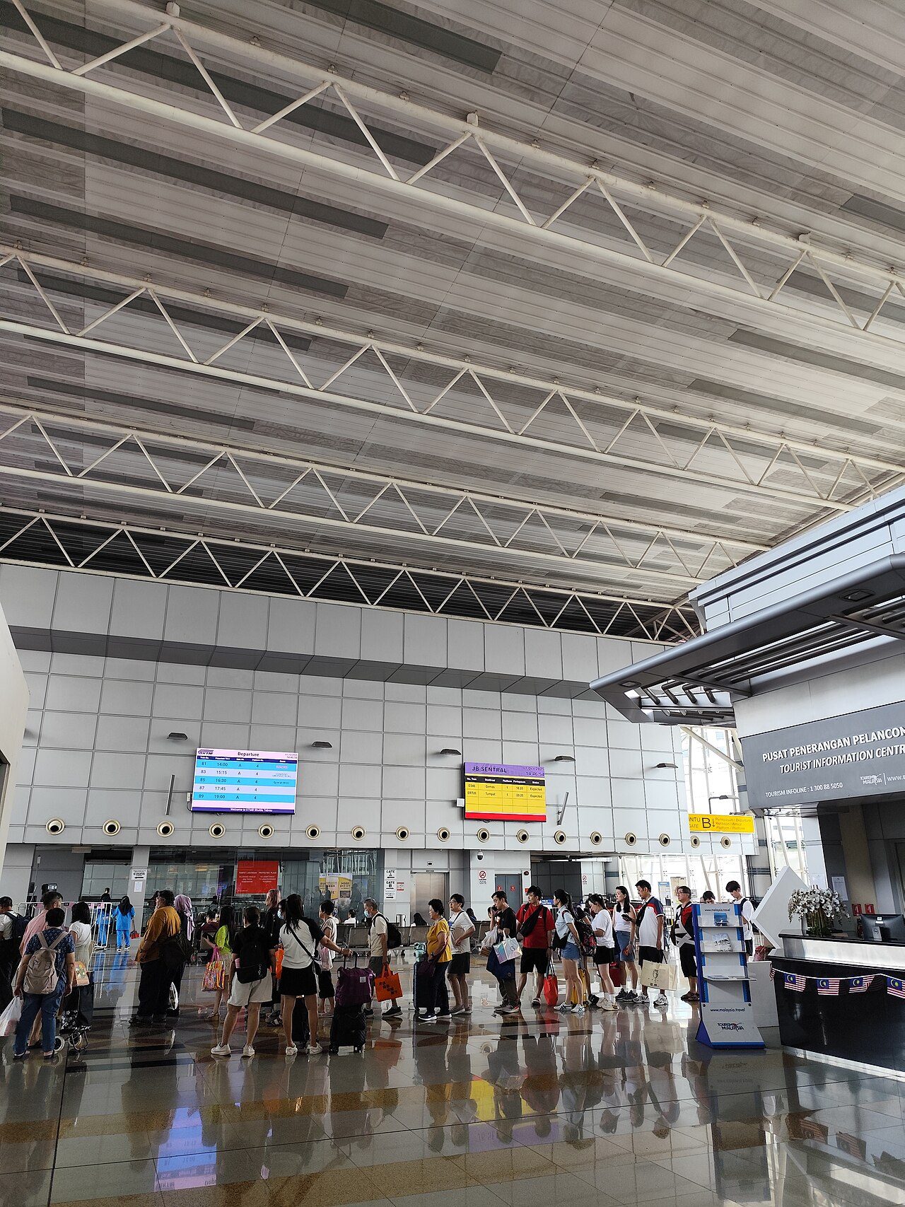 The concourse of JB Sentral station under a high white roof, with travellers queuing at the departure gates for the cross-border Shuttle Tebrau train