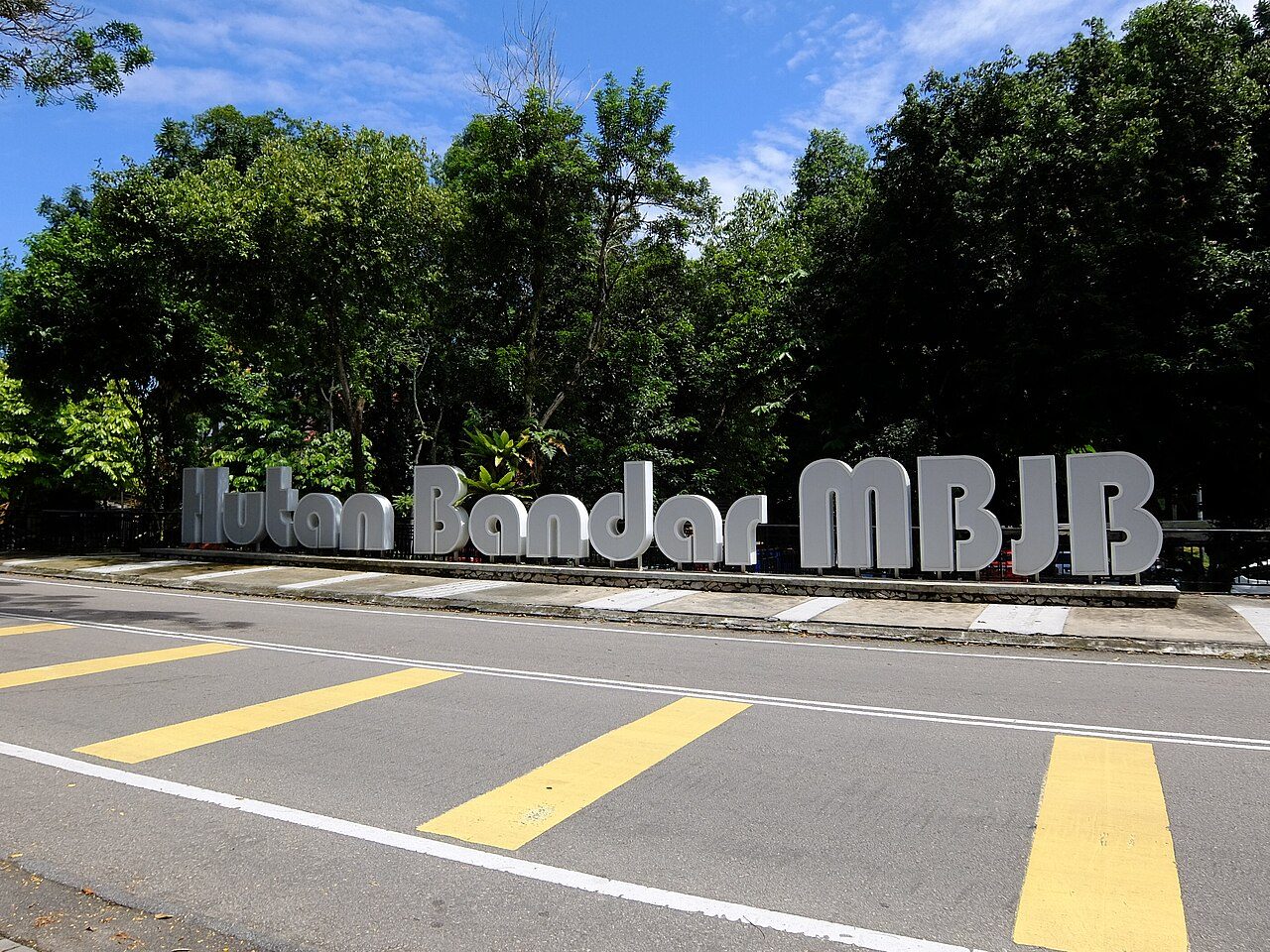Large white letters spelling Hutan Bandar MBJB in front of tall trees at the entrance to the Johor Bahru City Forest