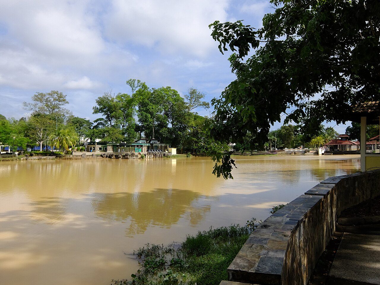 The brown lake at Taman Merdeka in Johor Bahru, with trees and park pavilions on the far bank
