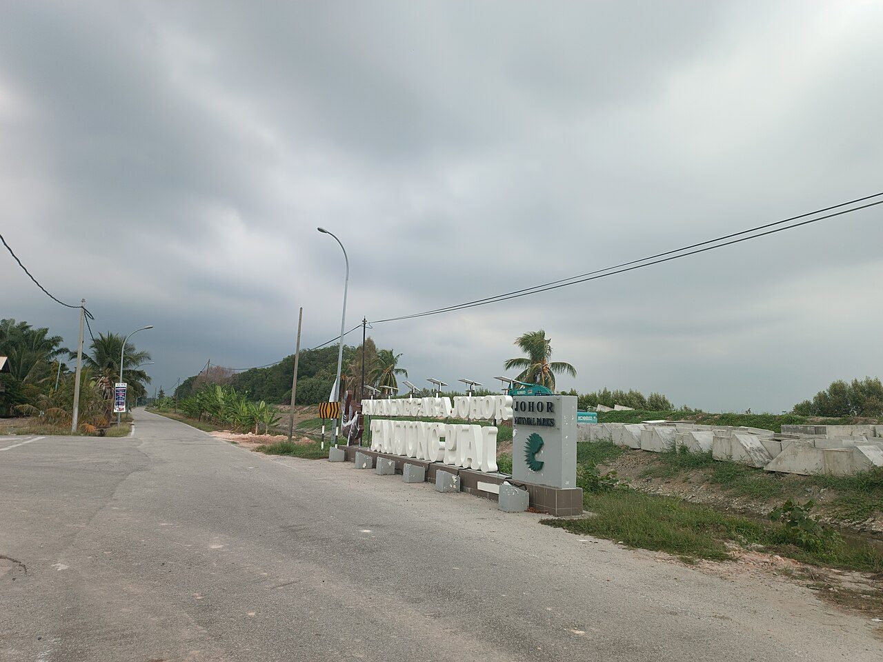 The Johor National Parks sign for Tanjung Piai beside the quiet approach road to the park under a cloudy sky