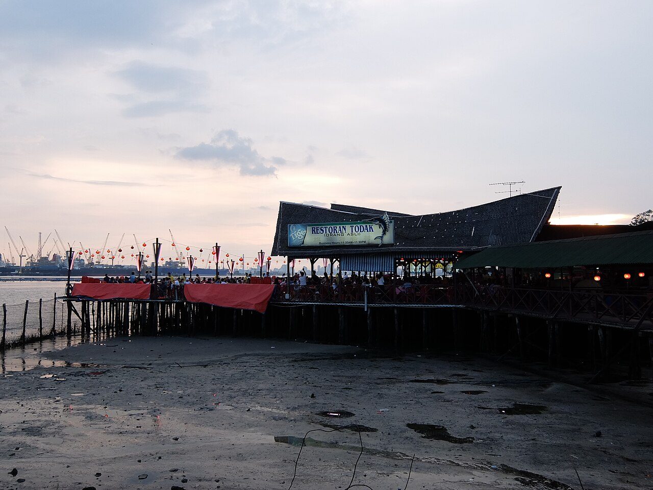 Restaurants in Johor Bahru: Restoran Todak, a seafood restaurant on wooden stilts over the water at Teluk Jawa, with port cranes on the skyline at dusk