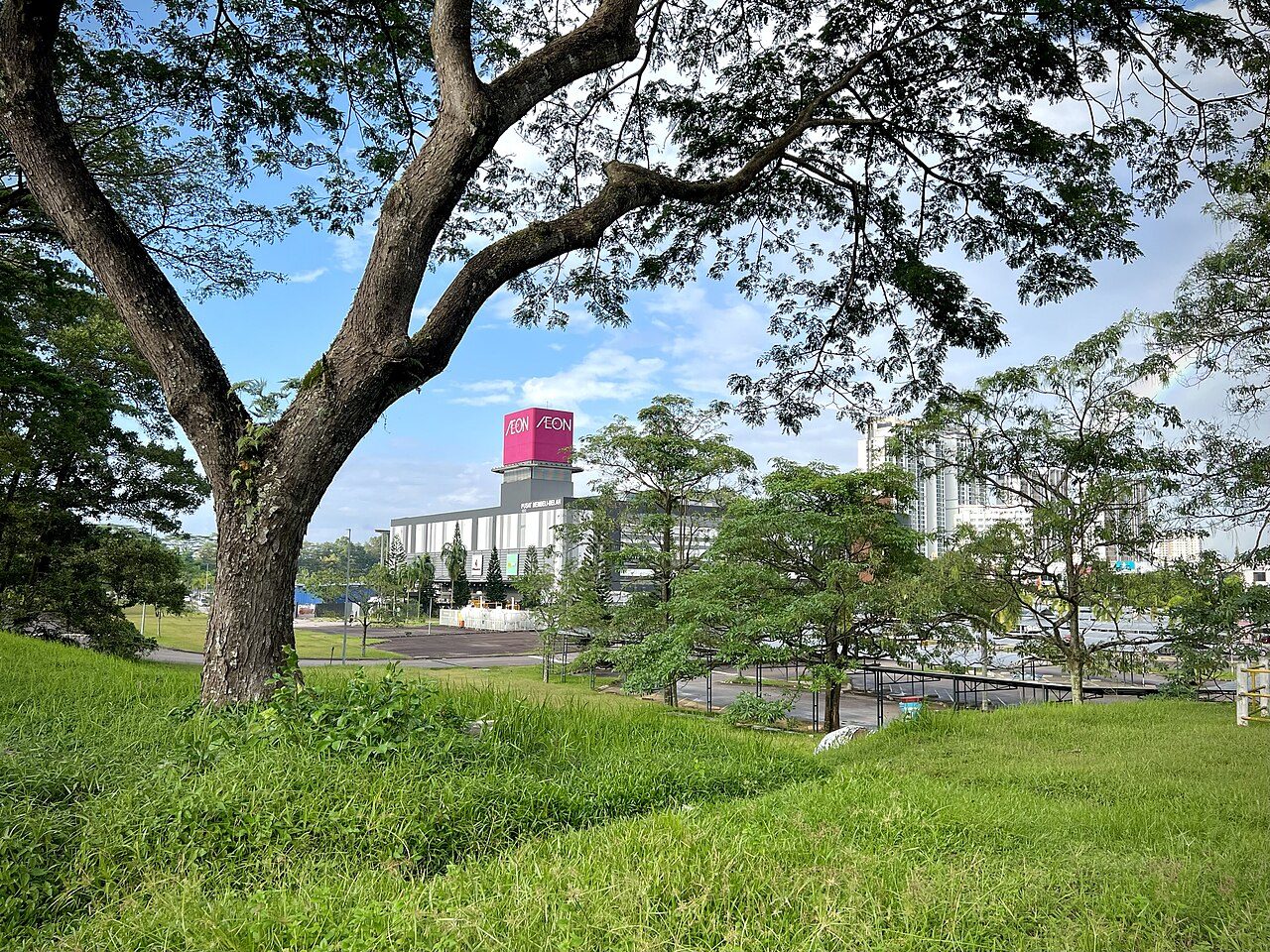 AEON Mall Bukit Indah seen across a grassy slope with large trees, its pink AEON tower visible behind the trees