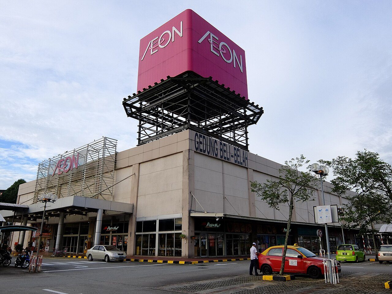 AEON Permas Jaya with its pink AEON sign tower above the grey mall building and a taxi parked in front