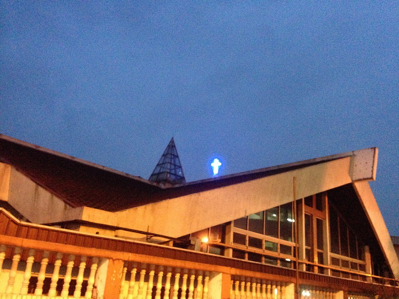 The sloping roof and glass spire of the Cathedral of the Sacred Heart in Johor Bahru at dusk, with a lit blue cross