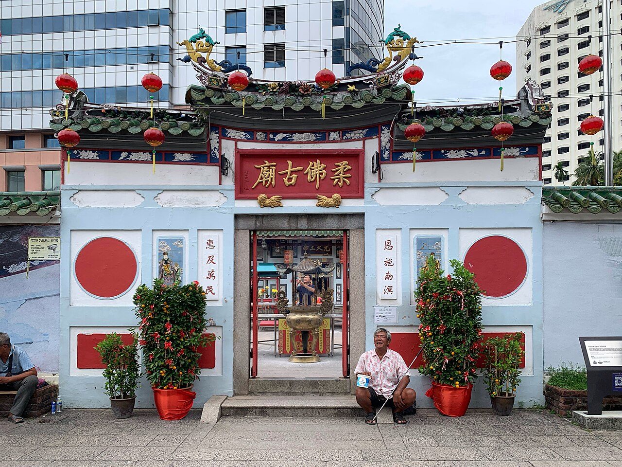 The front wall of the Johor Bahru Old Chinese Temple with red lanterns, a carved roof ridge and a gold-lettered name board, with office towers behind
