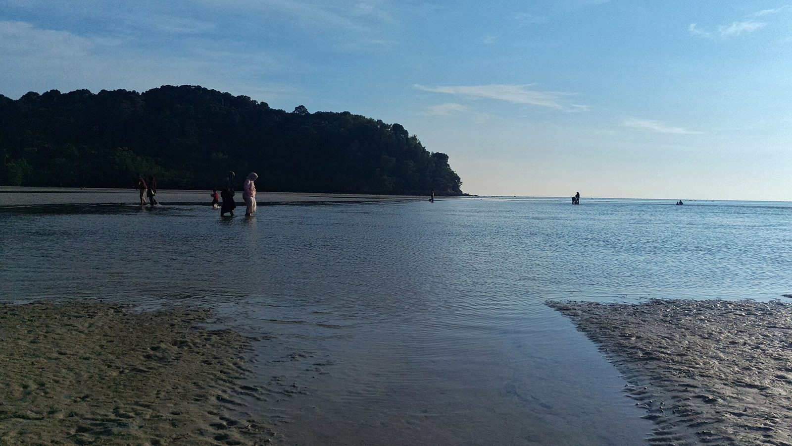 People wading across the shallow tide pools at Tanjung Biru (Blue Lagoon) beach with the forested Tanjung Tuan headland behind