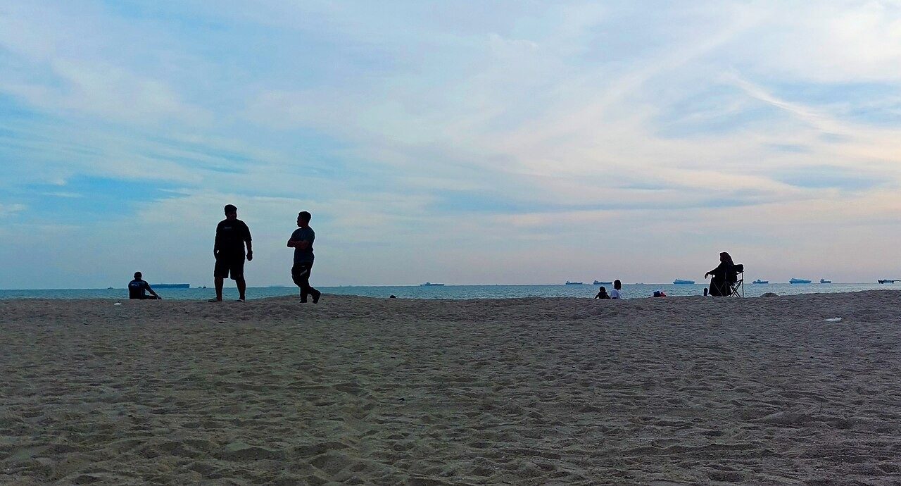 Melaka beaches and islands: people standing on the sand at Pantai Puteri at dusk with ships on the Strait of Malacca horizon
