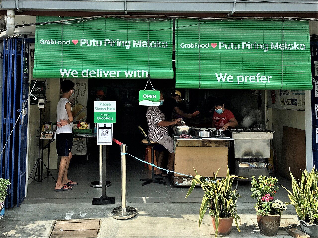Shopfront of Putu Piring Melaka on Jalan Tengkera with green blinds, a queue barrier and staff steaming putu piring, photographed in 2021