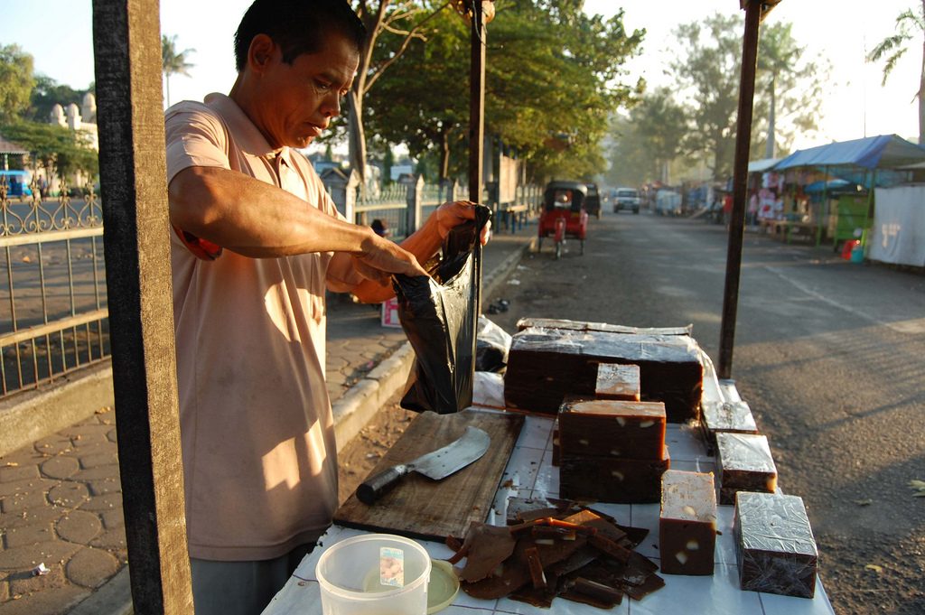 A vendor wrapping slices of dark brown dodol at a street stall in Indonesia, with more blocks on the table