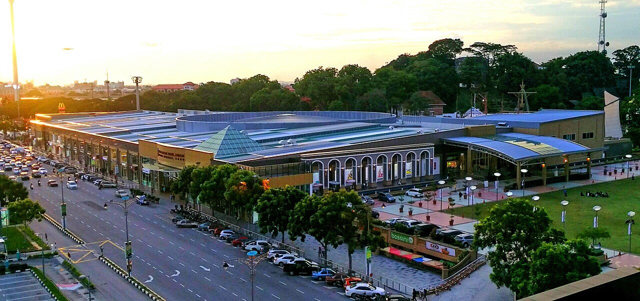 Dataran Pahlawan Melaka Megamall at sunset, a long low mall with a glass pyramid roof beside a lawn and a busy road