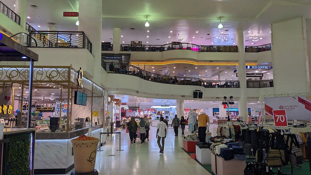The two-level main hall inside Mahkota Parade shopping centre, with shops, a drinks kiosk and shoppers walking through