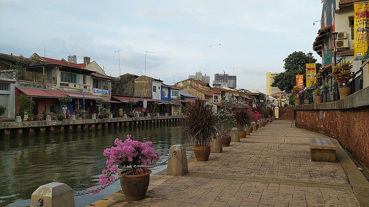 Paved riverside walkway with potted bougainvillea beside the Melaka River and old shophouses