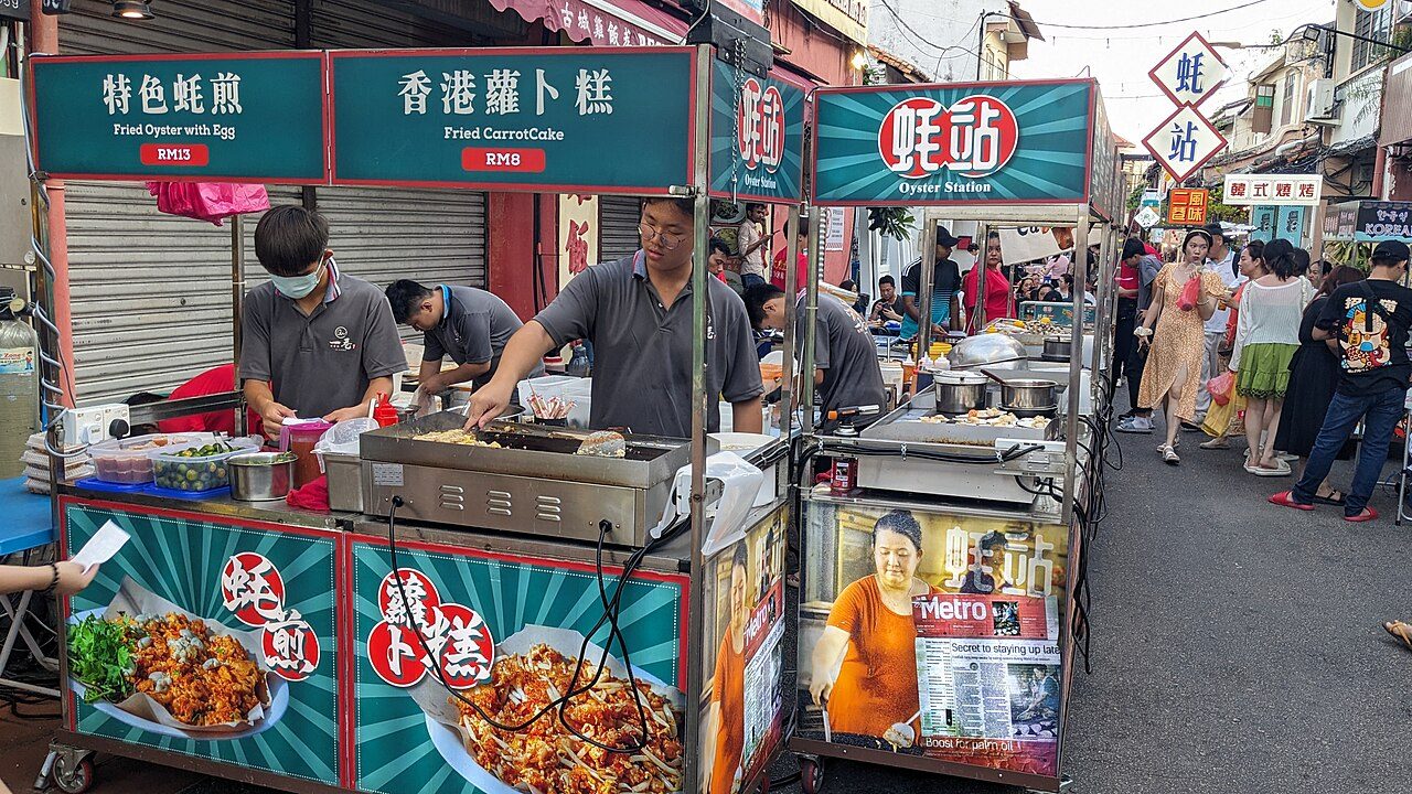 Fried oyster and fried carrot cake stalls with price boards at the Jonker Walk night market in Melaka, photographed in May 2024