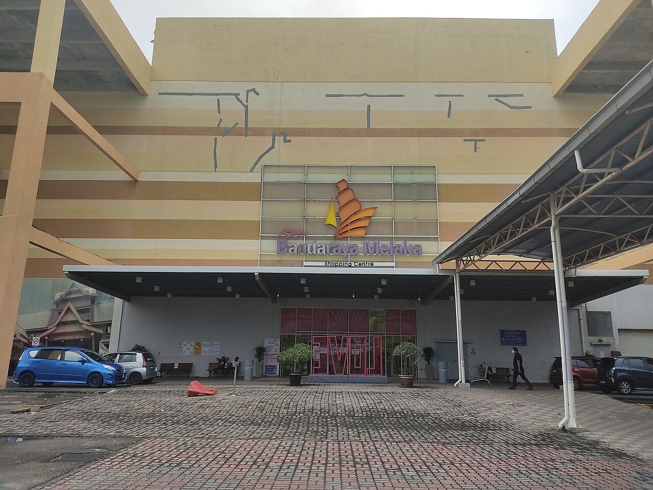 Front entrance of AEON Bandaraya Melaka Shopping Centre, with its ship logo above the doors and cars parked outside
