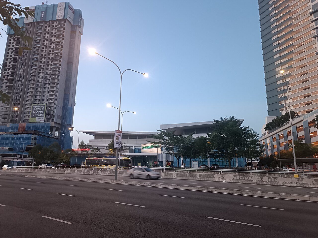 Columbia Asia Hospital Puchong at dusk, a low building with a 24-hour emergency sign, seen across a highway