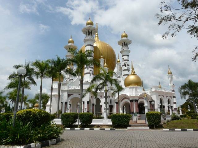White Ubudiah Mosque with golden domes and minarets behind palm trees and a paved forecourt