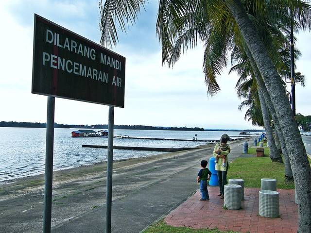 The palm-lined seafront promenade at Danga Bay in Johor Bahru beside the Strait of Johor, with a no-swimming sign in the foreground