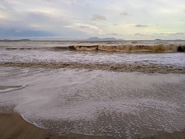 Brown, choppy waves breaking on the sand at Air Papan Beach near Mersing, with islands on the horizon