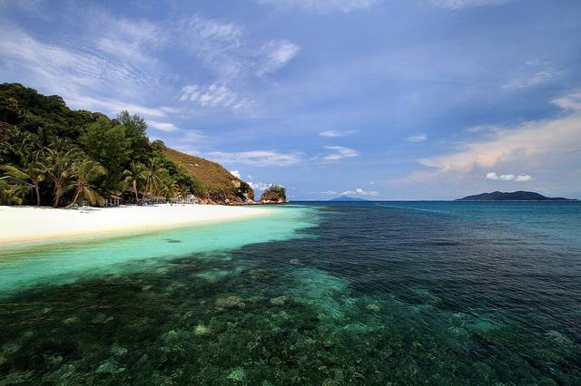 Coral patches in clear water off a white-sand beach with palms and a rocky headland at Pulau Rawa