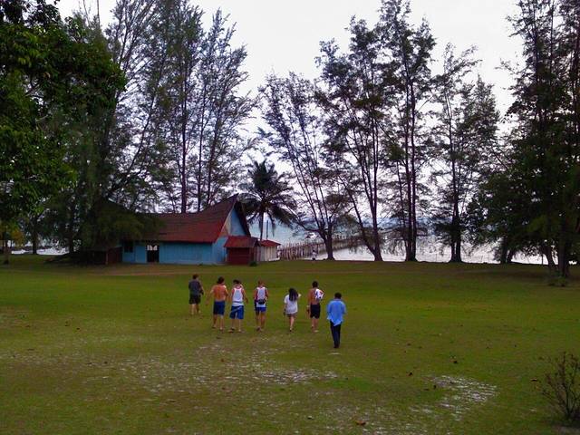 A group walking across a grassy clearing under casuarina trees towards a red-roofed building and jetty on Pulau Sibu