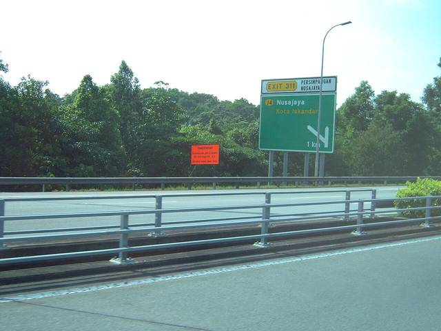 An expressway in Johor with a green exit sign for Nusajaya and Kota Iskandar beside a steel crash barrier
