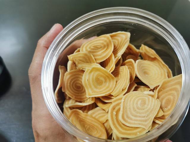 A hand holding a clear tub of kerepek bawang, thin spiral-shaped crackers sold in packets in Malaysia