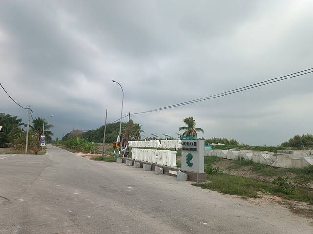 The Johor National Parks sign for Tanjung Piai beside the quiet approach road to the park under a cloudy sky