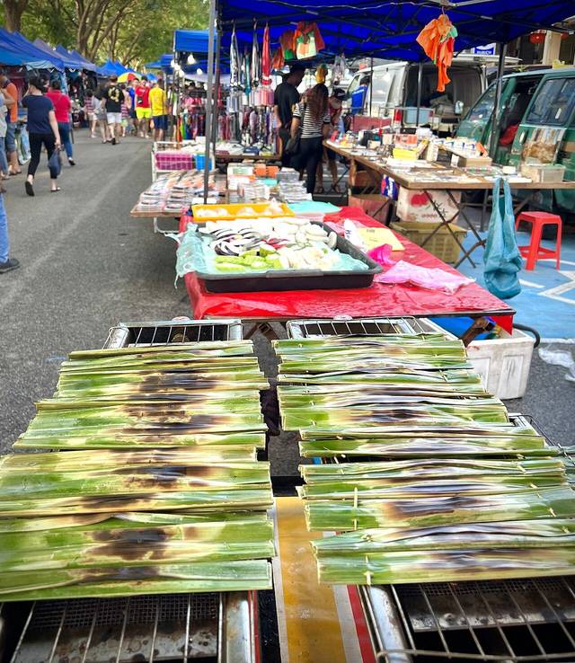 Otak-otak grilling in leaf wrappers at a stall in the Bukit Indah 2 Wednesday night market, with clothing and snack stalls behind, photographed in 2023