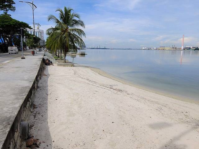 The sandy beach and palm-lined seafront road at Stulang Laut in Johor Bahru by day, looking along the Strait of Johor