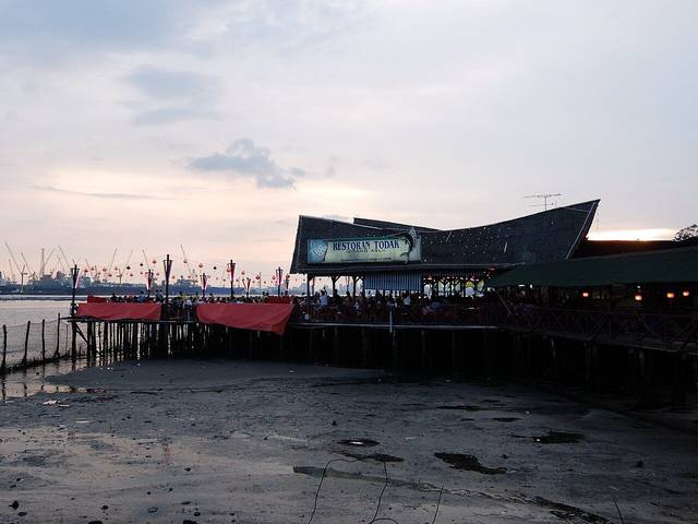 Restoran Todak, an open-sided seafood restaurant on wooden stilts over the mudflats at Teluk Jawa, Johor, with diners on the deck at dusk