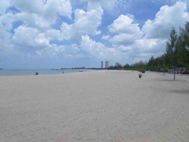 Wide, flat sand at Klebang Beach with casuarina trees and distant tower blocks under a blue sky