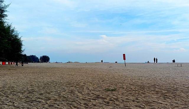 Looking south along the sand at Pantai Puteri, Tanjung Kling, with a few beachgoers and a red flag sign