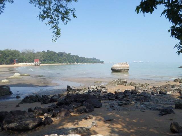 Low tide at Tanjung Bidara with dark rocks, a large boulder in the shallows and a wooded headland behind