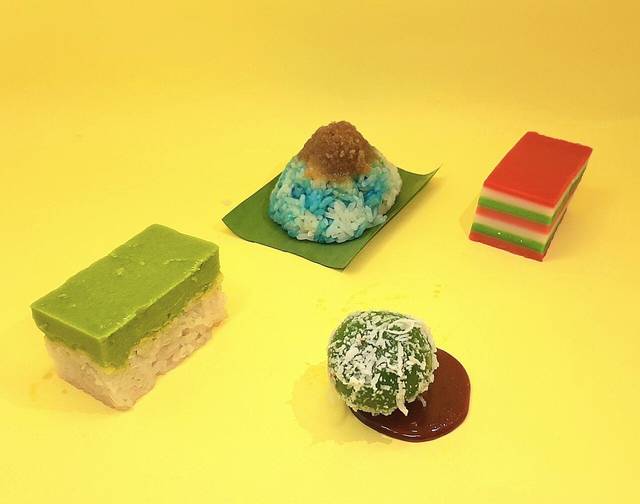 Four Nyonya kuih on a yellow table: pandan kaya on glutinous rice, blue pulut tai tai, layered kuih lapis and onde-onde, typical Melaka desserts