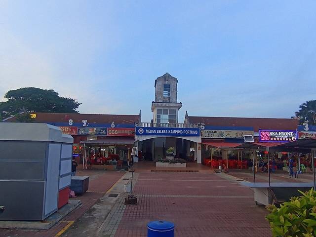Medan Selera Kampung Portugis in the Portuguese Settlement, Melaka, with numbered seafood restaurants either side of the central tower, photographed in June 2026